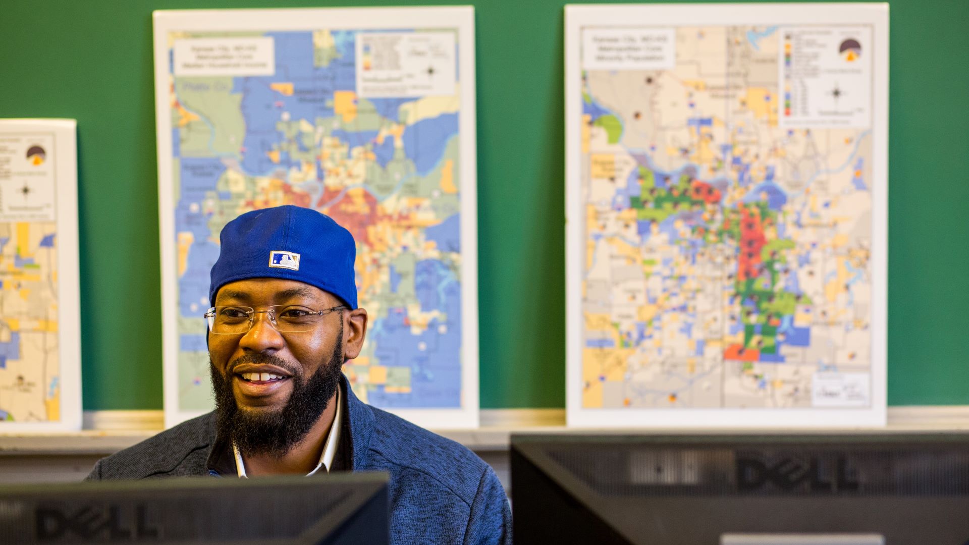 student wearing baseball cap sits at computer in front of maps
