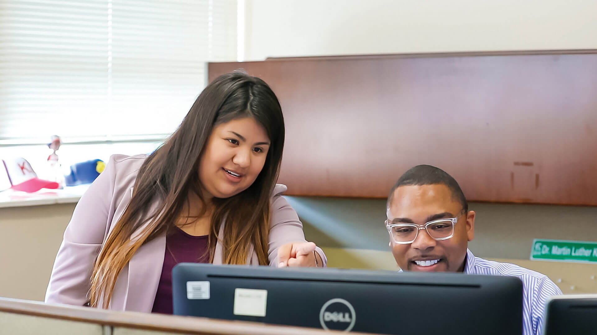 a mentor stands beside a student sitting at a computer and points out something on the monitor